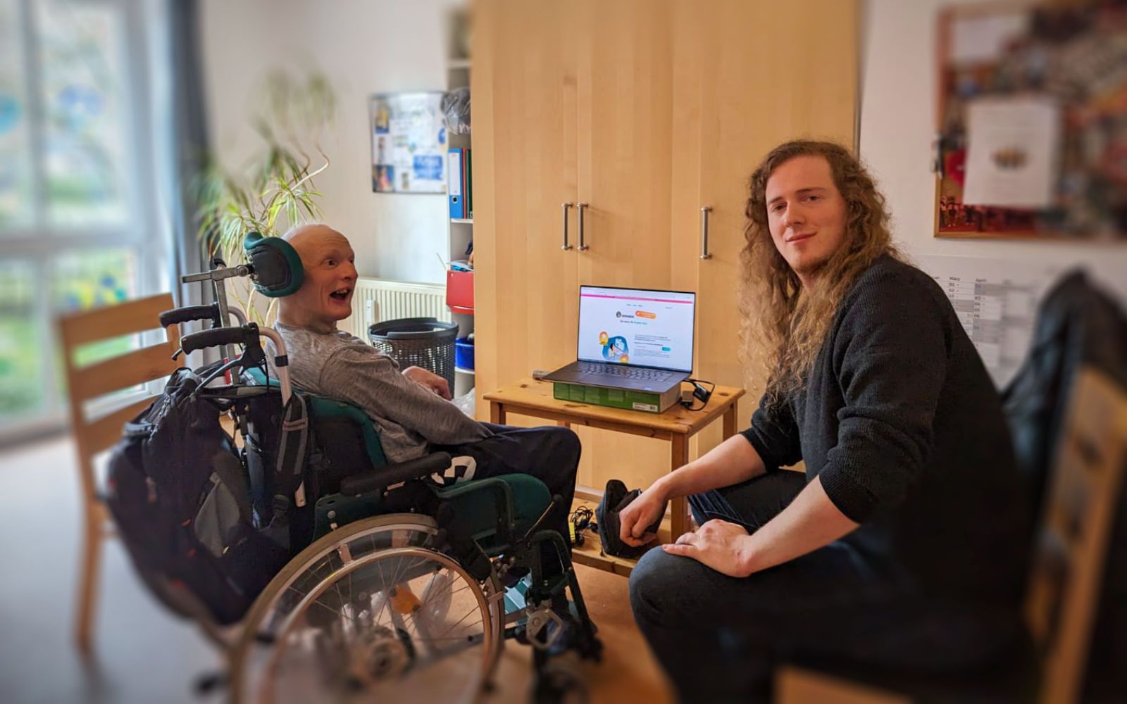 Links sitzt ein Proband im Rollstuhl, rechts sitzt Lukas auf einem Stuhl, in der Mitte ein Laptop mit Semanux.